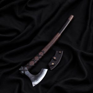 May include: A hand axe with a dark, weathered metal head and a long, straight wooden handle. The handle features a dark leather wrap in a crisscross pattern and a separate leather sheath with three metal snap buttons resting beside the axe head.