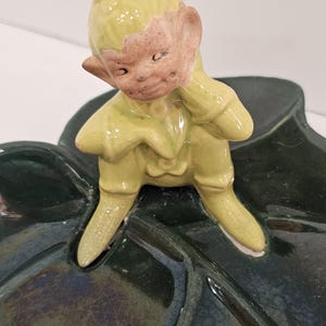 Puede incluir: Una figurita de cerámica de un duende verde sentado en una hoja verde. El duende lleva un sombrero verde y tiene la cara marrón.
