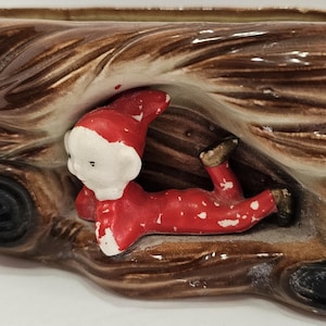 May include: A ceramic planter shaped like a tree trunk with a small elf figurine lying inside. The elf is wearing a red and white suit with a pointed hat.