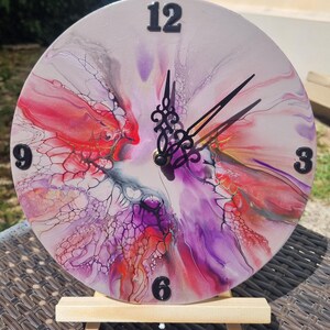 May include: A round decorative clock with a colorful abstract design in shades of red, purple, and white. Black numbers mark the hours, and black hands indicate the time. The clock is displayed on a small wooden easel.