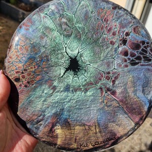 Peut inclure: Une peinture abstraite circulaire avec un vide noir central, entouré d'un réseau de teintes turquoise, violettes et dorées. L'œuvre présente un motif fluide en forme de cellules et est signée "Kat's Colors".