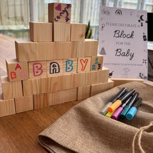 May include: A wooden block tower with the word "BABY" spelled out in colourful letters. The blocks are decorated with markers and numbers. A sign reads "Please decorate a block for the baby. Don't forget to sign your name."  A hessian bag with colourful markers is in the foreground.