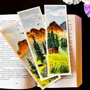 May include: Watercolor landscape bookmarks featuring mountain scenes with trees, a cabin, and birds. The bookmarks are hand-painted with vibrant colors and are displayed on an open book.