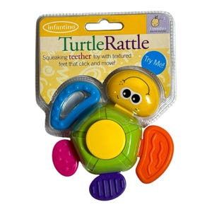 May include: A Turtle Rattle baby toy in a clear plastic package. The toy is green, yellow, blue, orange, purple, and pink. The text on the package says "Turtle Rattle" and "Squeaking teether toy with textured feet that click and move!"