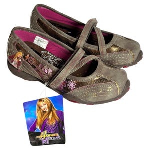 Puede incluir: Par de zapatos grises y marrones estilo Mary Jane con cierre de correa. Los zapatos presentan un interior rosa, detalles florales y de notas musicales, y una etiqueta con el logo de Hannah Montana.
