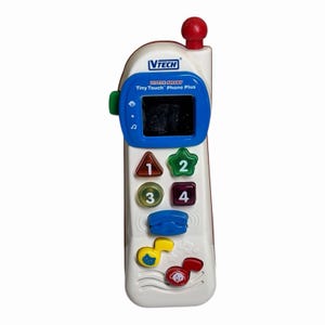 May include: A white and red VTech Little Smart Tiny Touch Phone Plus toy phone. The toy has a blue screen, colourful buttons with numbers and shapes, and a red antenna. The phone has musical notes and a dog graphic.