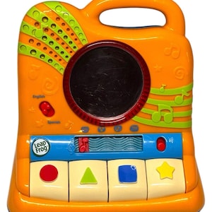 Puede incluir: Juguete musical LeapFrog naranja y verde con asa. Cuenta con una pantalla negra, botones y un teclado con formas. Incluye las palabras "English" y "Spanish".