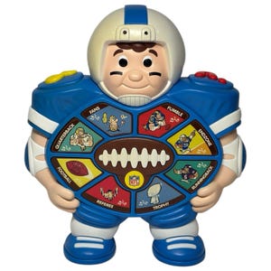 May include: A vintage football-themed toy in the shape of a cartoon character wearing a helmet and jersey. The toy is primarily blue, white, and tan, with a dial featuring football-related icons and the NFL logo. The toy has buttons and is designed for children.