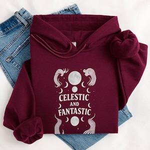 Op de afbeelding: Een bordeauxrode hoodie met een hemels ontwerp met manen en sterren, en de woorden "Celestial and Fantastic" in het wit. De hoodie is opgevouwen op een blauwe jeans.