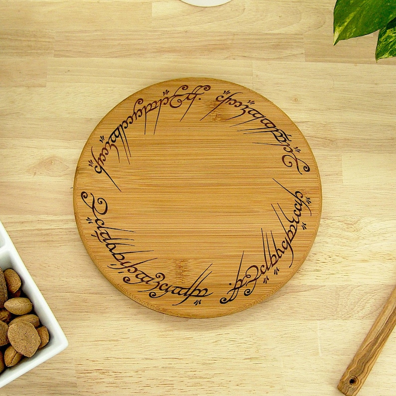 K&ouml;nnte beinhalten: Rundes Servierbrett aus Holz mit eingravierter Schrift am Rand. Das Brett ist aus hellem Holz gefertigt und hat eine nat&uuml;rliche Maserung. Die eingravierte Schrift ist dunkel. Das Brett liegt auf einer Holzoberfl&auml;che.