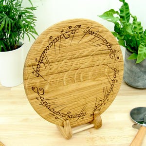 Puede incluir: Una tabla de cortar redonda de madera con un acabado marrón claro. La tabla presenta una inscripción grabada en un patrón circular. Se muestra sobre un pequeño soporte de madera, con un cortador de pizza visible en la esquina. La tabla tiene unos 25 cm de diámetro.