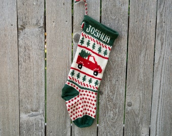 May include: A green and white Christmas stocking with a red truck carrying a Christmas tree. The stocking has the name "Joshua" embroidered on it.