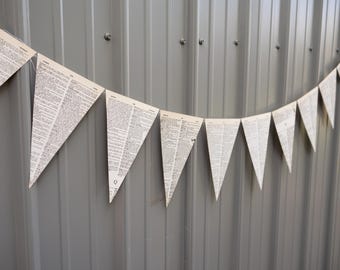 May include: A string of triangular paper pennants, each printed with text, hangs against a gray corrugated metal wall. The pennants are strung together, creating a decorative banner. The paper has a vintage, aged appearance.