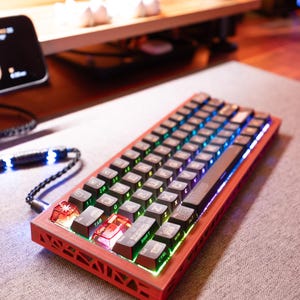 Puede incluir: Un teclado mecánico rojo y negro con un conjunto de teclas personalizadas. El teclado tiene una variedad de teclas, incluidas teclas de función, teclas de flecha y teclas numéricas. El teclado está iluminado con iluminación RGB.