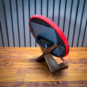 Puede incluir: Un objeto redondo rojo y negro con un asa, apoyado en un soporte de madera. El soporte tiene un dise&ntilde;o en forma de X. El fondo presenta un patr&oacute;n de rayas y una superficie de madera.