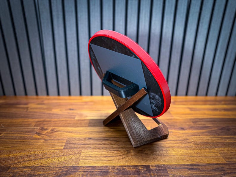Puede incluir: Un objeto redondo rojo y negro con un asa, apoyado en un soporte de madera. El soporte tiene un dise&ntilde;o en forma de X. El fondo presenta un patr&oacute;n de rayas y una superficie de madera.