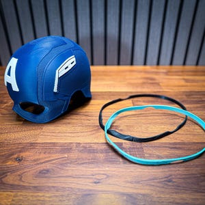 May include: A blue Captain America helmet with a white "A" on the side. The helmet has a textured surface and is accompanied by a black and a turquoise headband. The items are on a wooden surface.