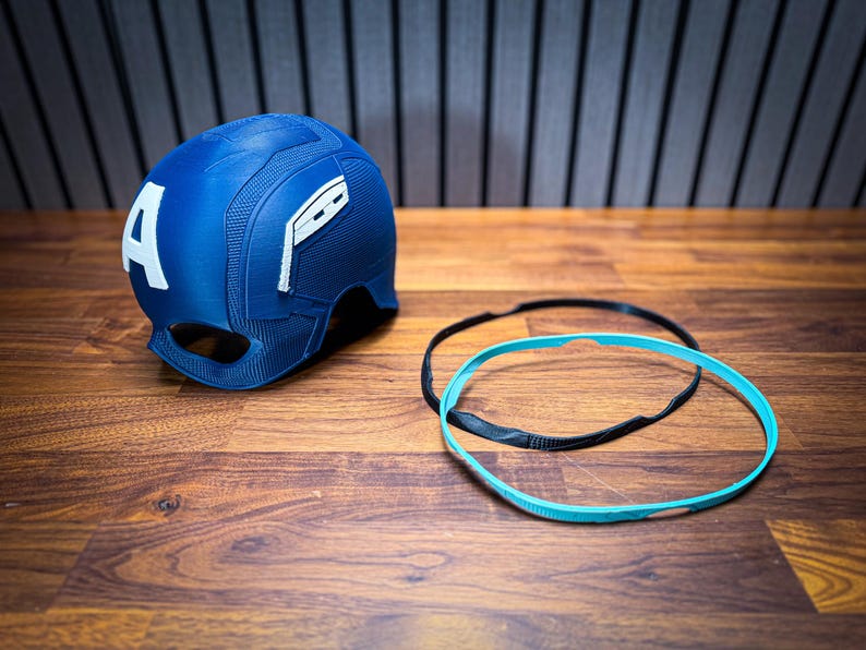 May include: A blue Captain America helmet with a white "A" on the side. The helmet has a textured surface and is accompanied by a black and a turquoise headband. The items are on a wooden surface.