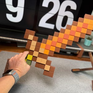 May include: A pixelated sword in shades of brown, orange, and yellow, reminiscent of a video game design. The sword is held in a hand, with a gray watch visible on the wrist. The background includes a digital clock displaying the time.