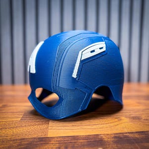 May include: A blue helmet with white accents, reminiscent of a superhero's attire, sits on a wooden surface. The helmet features the letter "A" on the side and a textured design. The overall aesthetic suggests a cosplay or collectible item.