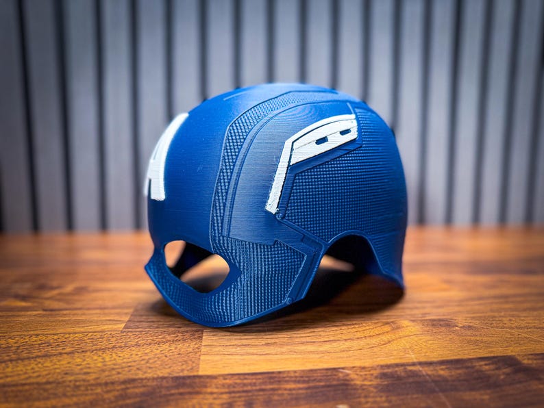 May include: A blue helmet with white accents, reminiscent of a superhero's attire, sits on a wooden surface. The helmet features the letter "A" on the side and a textured design. The overall aesthetic suggests a cosplay or collectible item.