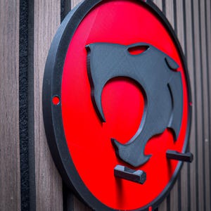 Puede incluir: Decoración mural redonda con borde negro, fondo rojo y silueta de cabeza de pantera negra. Tiene dos clavijas de montaje negras. La obra de arte está montada sobre una superficie de madera con lamas verticales.