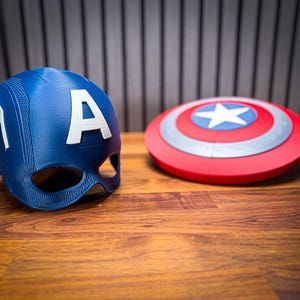 May include: A blue Captain America helmet with a white "A" and a red, white, and blue shield. The shield has a white star in the center. Both items are on a wooden surface.