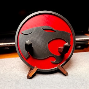May include: A round, red and black decorative item featuring a stylized panther head design. The item is mounted on a wooden stand. The panther head is black, and the background is red.