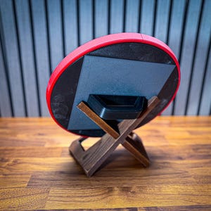 Puede incluir: Un objeto circular rojo y negro, posiblemente un altavoz o una pieza decorativa, est&aacute; apoyado en un soporte de madera. El soporte tiene una base en forma de X y un acabado marr&oacute;n oscuro. El fondo presenta una superficie de madera y una pared con l&iacute;neas verticales.
