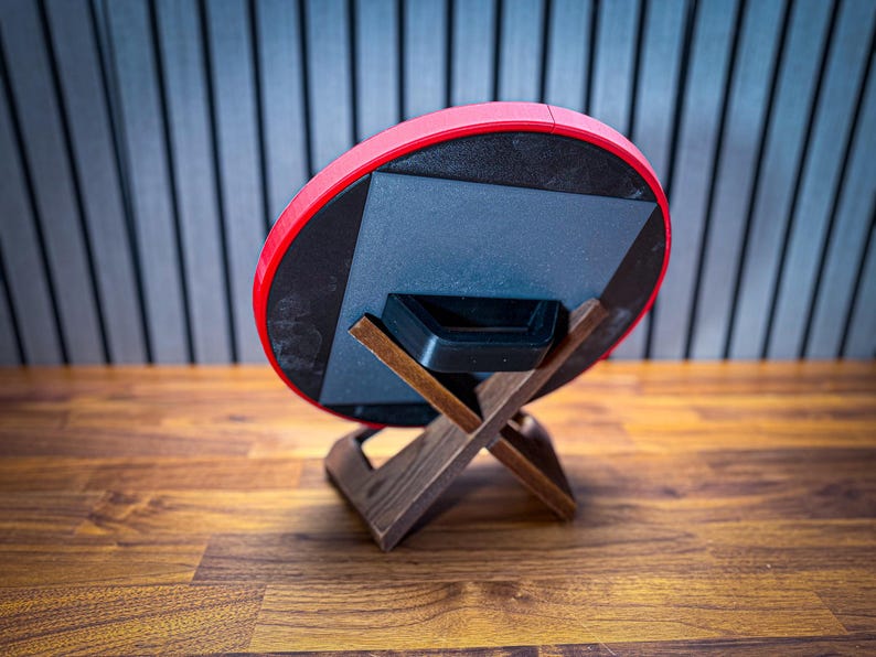 Puede incluir: Un objeto circular rojo y negro, posiblemente un altavoz o una pieza decorativa, est&aacute; apoyado en un soporte de madera. El soporte tiene una base en forma de X y un acabado marr&oacute;n oscuro. El fondo presenta una superficie de madera y una pared con l&iacute;neas verticales.