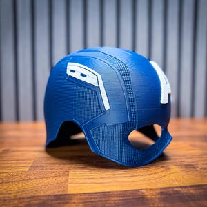 May include: A blue and white Captain America helmet, a replica of the iconic superhero's headgear. The helmet features a textured design and the letter "A" on the side. It is made of a hard material and sits on a wooden surface.