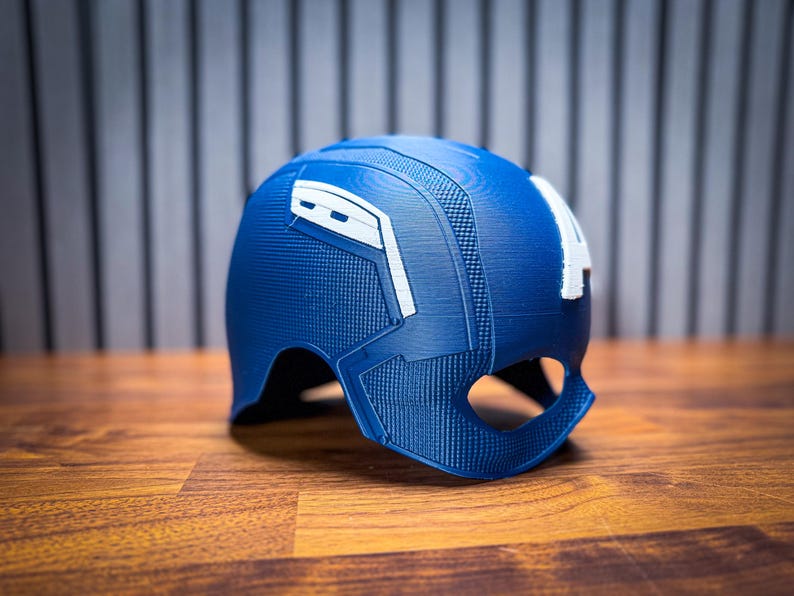 May include: A blue and white Captain America helmet, a replica of the iconic superhero's headgear. The helmet features a textured design and the letter "A" on the side. It is made of a hard material and sits on a wooden surface.