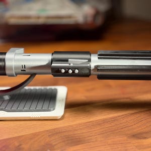 May include: A silver and black lightsaber with a red blade, on a white and grey stand. The hilt has a ribbed design and three small buttons. The lightsaber is a prop from a science fiction franchise.