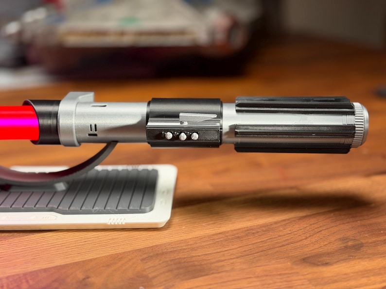 May include: A silver and black lightsaber with a red blade, on a white and grey stand. The hilt has a ribbed design and three small buttons. The lightsaber is a prop from a science fiction franchise.