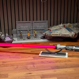 May include: A red lightsaber with a silver and black handle rests on a white stand. In the background are Star Wars themed models, including a Millennium Falcon and a model of Darth Vader's ship.