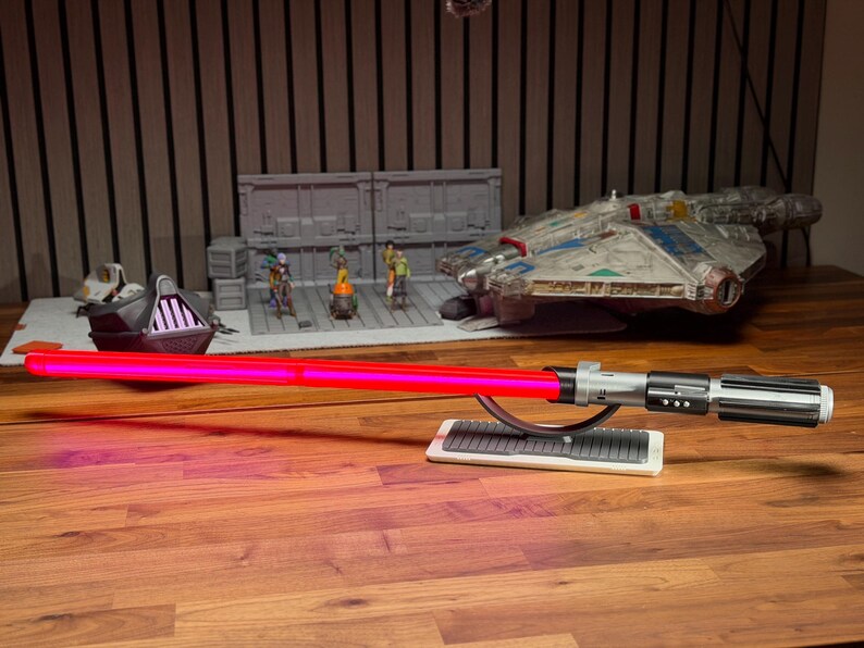 May include: A red lightsaber with a silver and black handle rests on a white stand. In the background are Star Wars themed models, including a Millennium Falcon and a model of Darth Vader's ship.