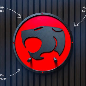 May include: A multi-color sword holder featuring a black silhouette of a cat-like creature on a red circular background, framed by a black border. The design is mounted on a dark wood-grain surface.