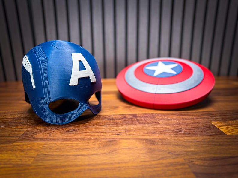 Puede incluir: Un casco azul de Capit&aacute;n Am&eacute;rica con una "A" blanca y un escudo rojo, blanco y azul con una estrella blanca. Los objetos est&aacute;n sobre una superficie de madera.