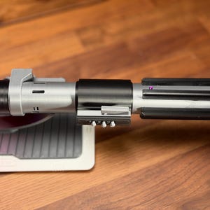 May include: A detailed replica lightsaber with a red blade and a metallic silver and black hilt. The hilt has intricate details, including a ribbed section and various buttons. The lightsaber rests on a grey and white stand.