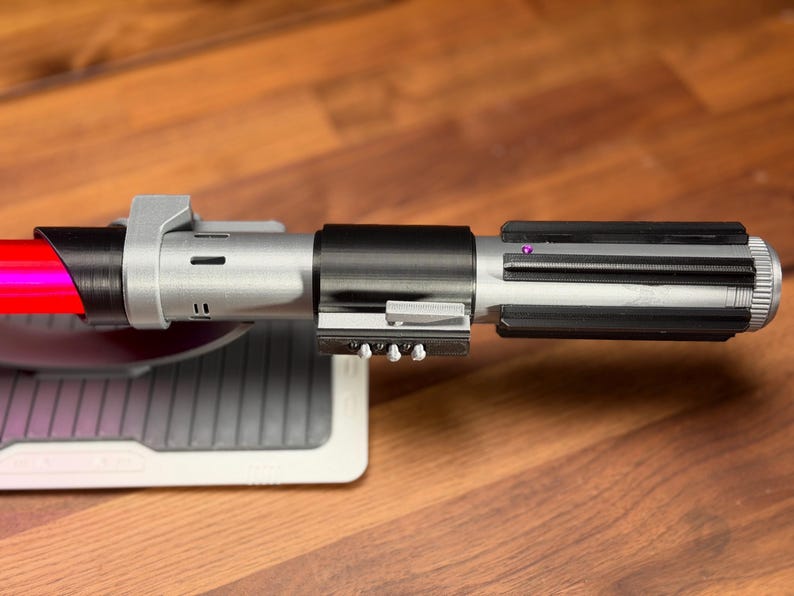 May include: A detailed replica lightsaber with a red blade and a metallic silver and black hilt. The hilt has intricate details, including a ribbed section and various buttons. The lightsaber rests on a grey and white stand.