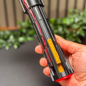 Op de afbeelding: Een zwart en rood lichtzwaard rekwisiet met gouden en zilveren accenten. De prop heeft rode bedrading en een rode knop aan de basis. Het lichtzwaard wordt in een hand gehouden, met een vage groene achtergrond.
