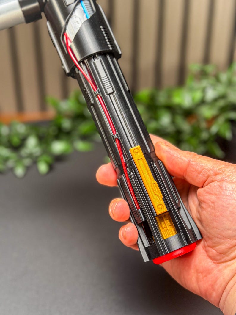 Op de afbeelding: Een zwart en rood lichtzwaard rekwisiet met gouden en zilveren accenten. De prop heeft rode bedrading en een rode knop aan de basis. Het lichtzwaard wordt in een hand gehouden, met een vage groene achtergrond.