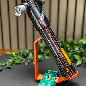 Op de afbeelding: Een gedetailleerd zwart lichtzwaard rekwisiet met zilveren en gekleurde accenten, tentoongesteld op een oranje standaard met groene accessoires. Het lichtzwaard heeft een kruisbeschermingsontwerp en is ideaal voor cosplay of displaydoeleinden.