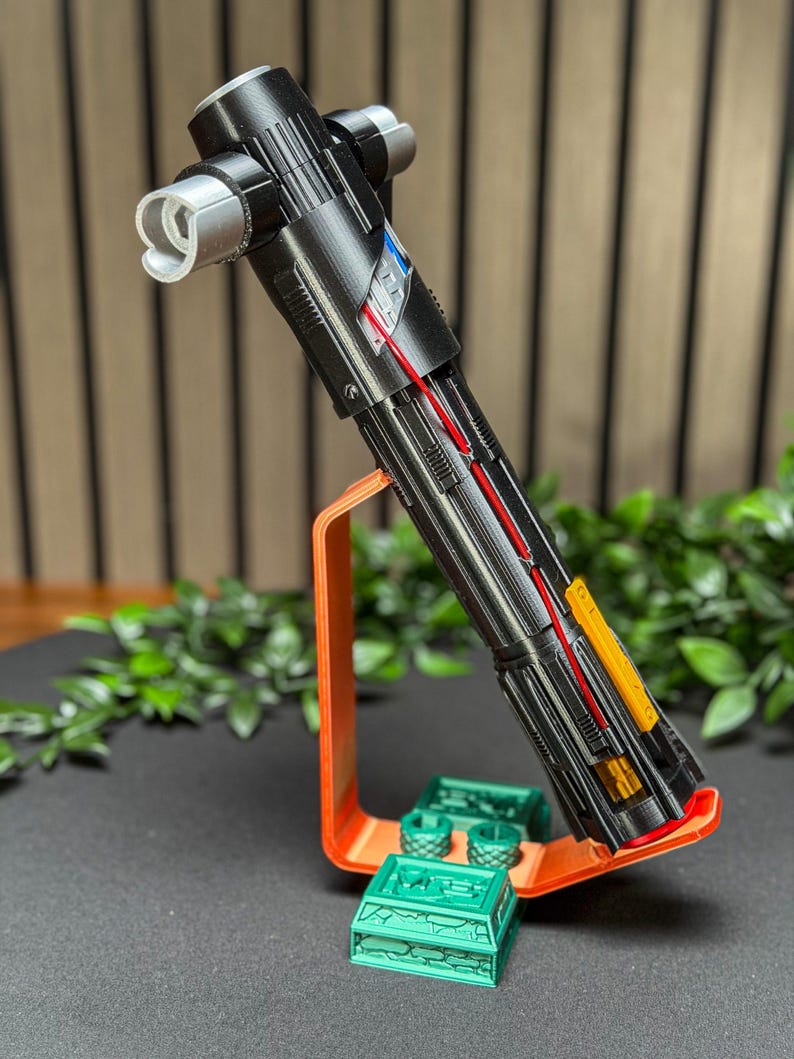 Op de afbeelding: Een gedetailleerd zwart lichtzwaard rekwisiet met zilveren en gekleurde accenten, tentoongesteld op een oranje standaard met groene accessoires. Het lichtzwaard heeft een kruisbeschermingsontwerp en is ideaal voor cosplay of displaydoeleinden.