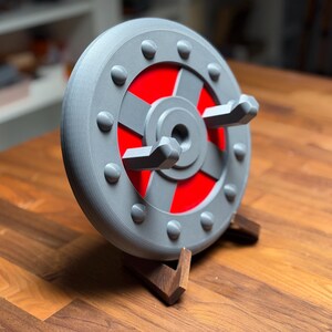 Puede incluir: Una rueda decorativa gris y roja con un cubo central y asas salientes. La rueda está montada sobre un soporte de madera. El diseño presenta una serie de remaches grises alrededor del borde exterior y un centro rojo.