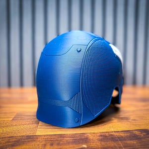 May include: A blue helmet with a textured surface and a white accent. The helmet has a detailed design with rivets and panels. The object is on a wooden surface.