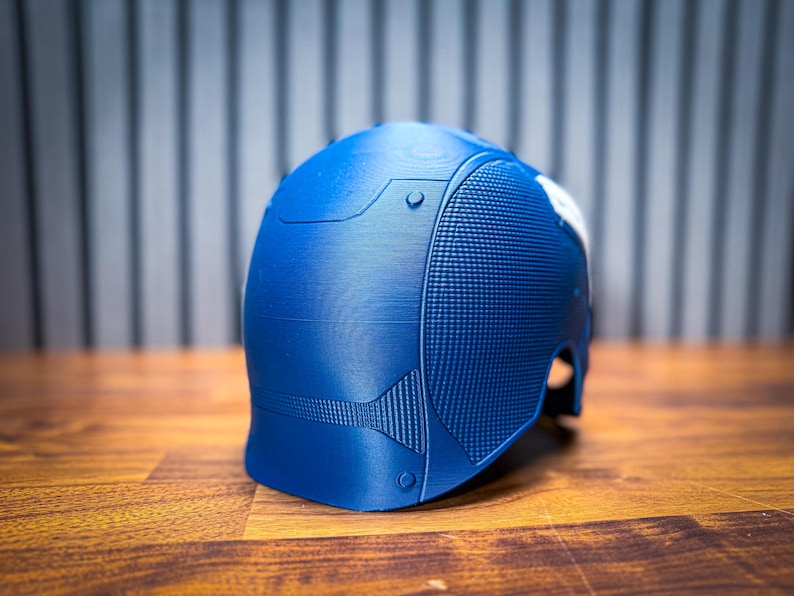 May include: A blue helmet with a textured surface and a white accent. The helmet has a detailed design with rivets and panels. The object is on a wooden surface.