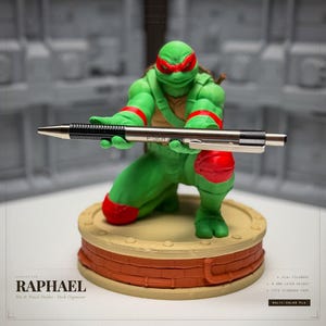 May include: A green Raphael figurine, a Teenage Mutant Ninja Turtle, holding a silver pen. The figure is on a round, tan and brown base. The pen has a black grip and the text "F301" on it. The base is labeled "RAPHAEL Pen & Pencil Holder - Desk Organizer."
