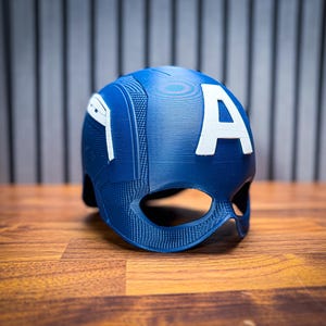 May include: A blue helmet with a white "A" on the back, reminiscent of a superhero's attire. The helmet has a textured surface and eye openings. The item is placed on a wooden surface.