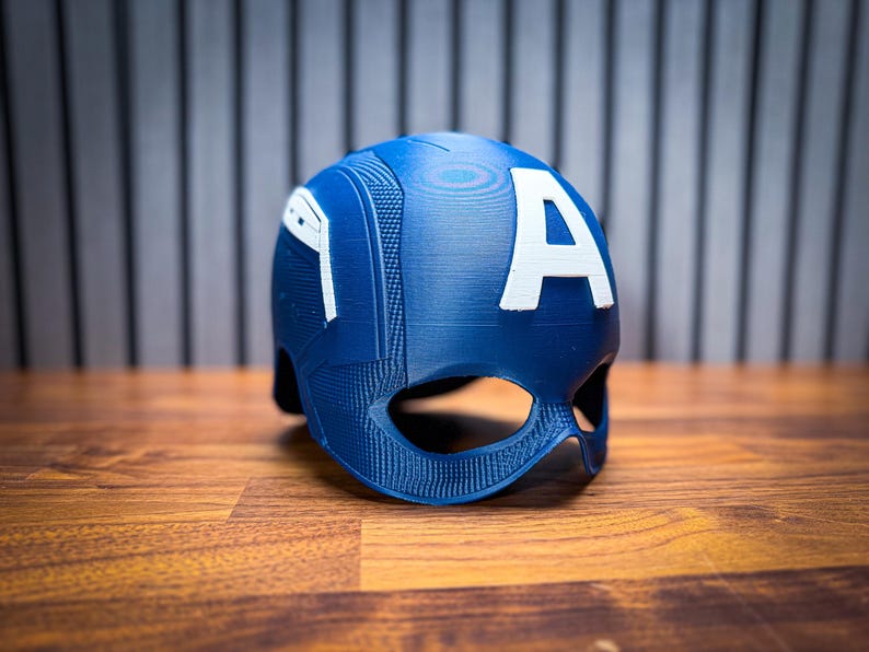 May include: A blue helmet with a white "A" on the back, reminiscent of a superhero's attire. The helmet has a textured surface and eye openings. The item is placed on a wooden surface.